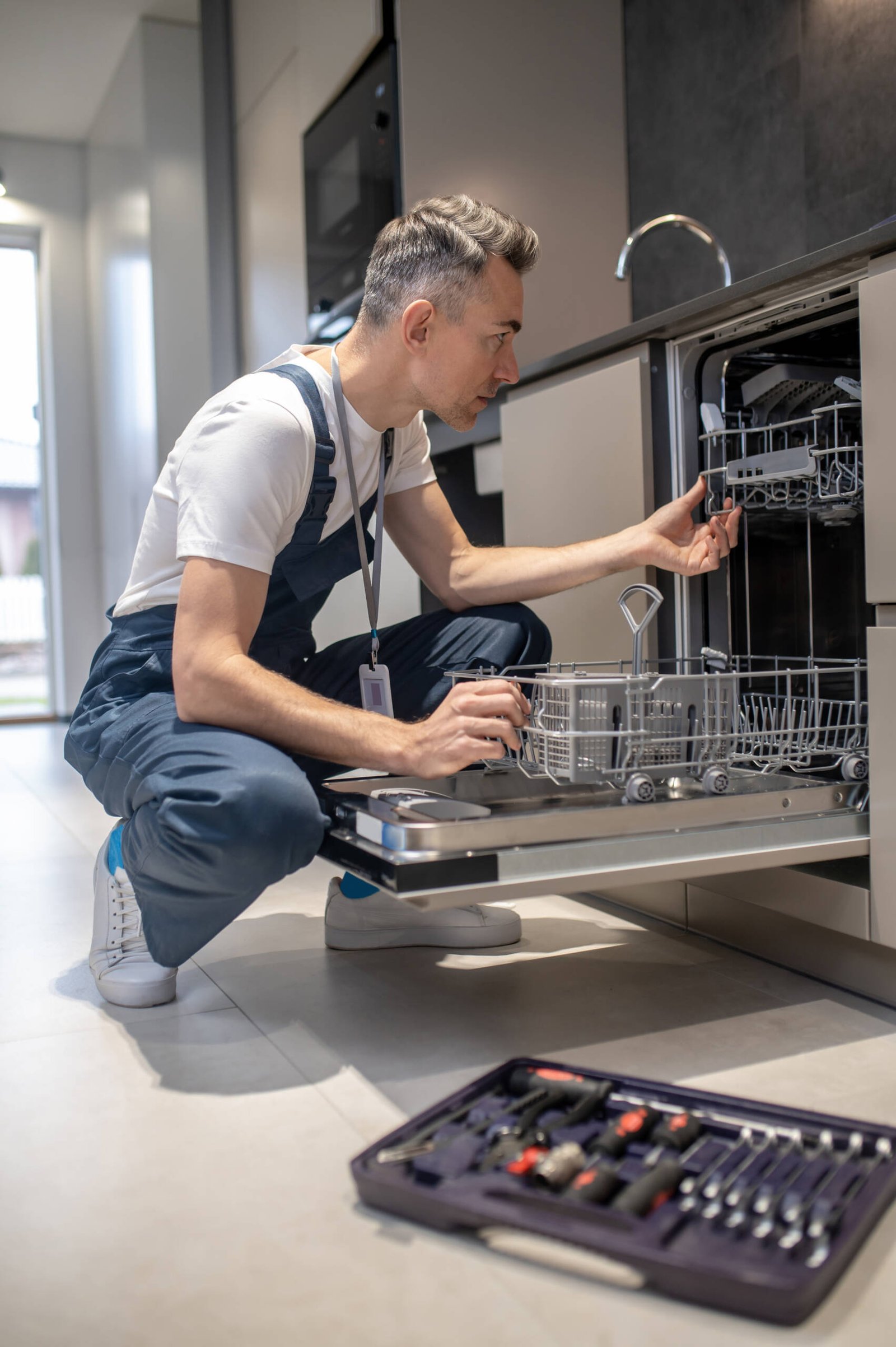 Diagnostics of equipment. Profile of serious man in dark overalls with badge crouching attentively looking at open dishwasher in kitchen at home during day