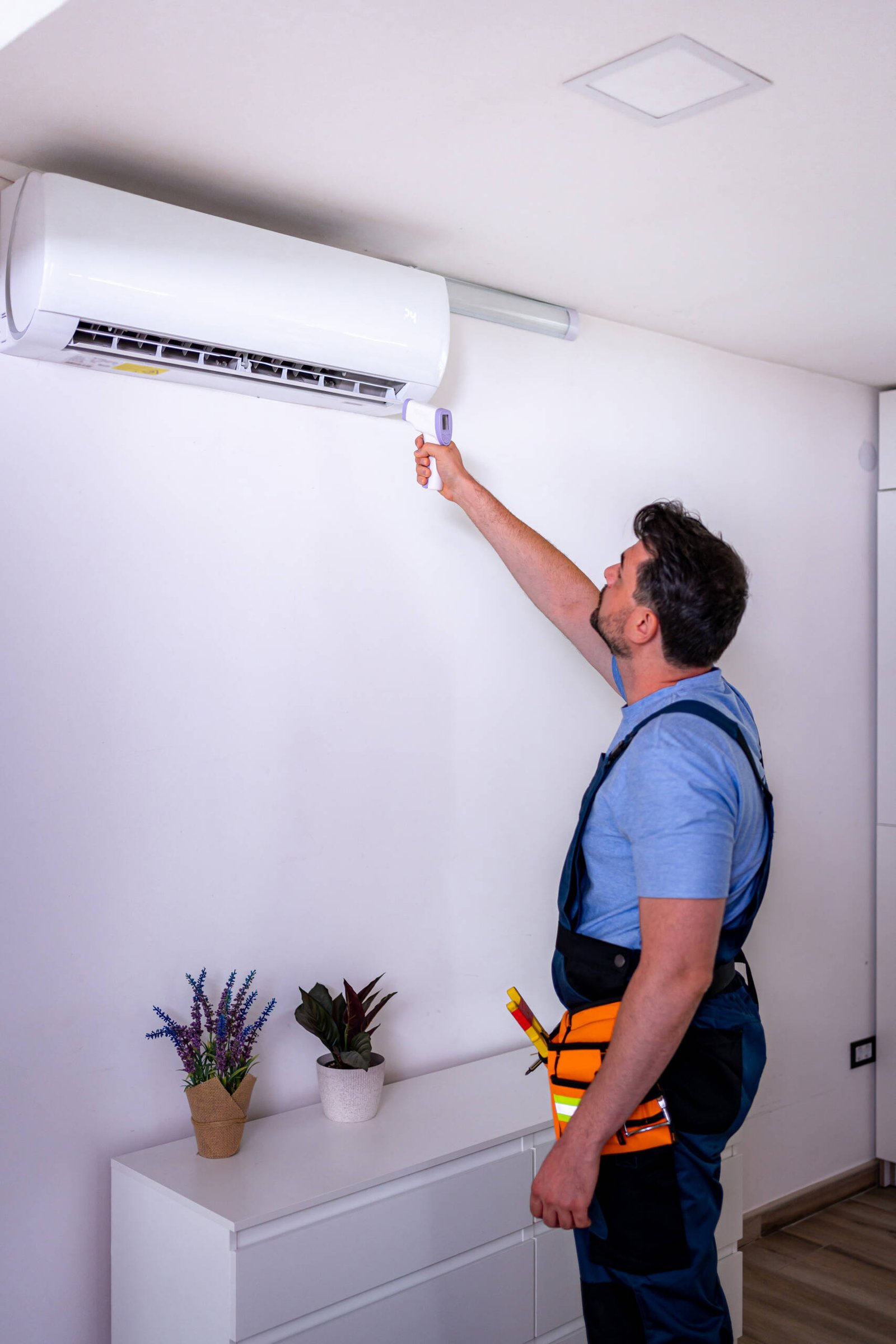 A worker uses a remote control to check the air conditioning unit on the wall in a room with plants.